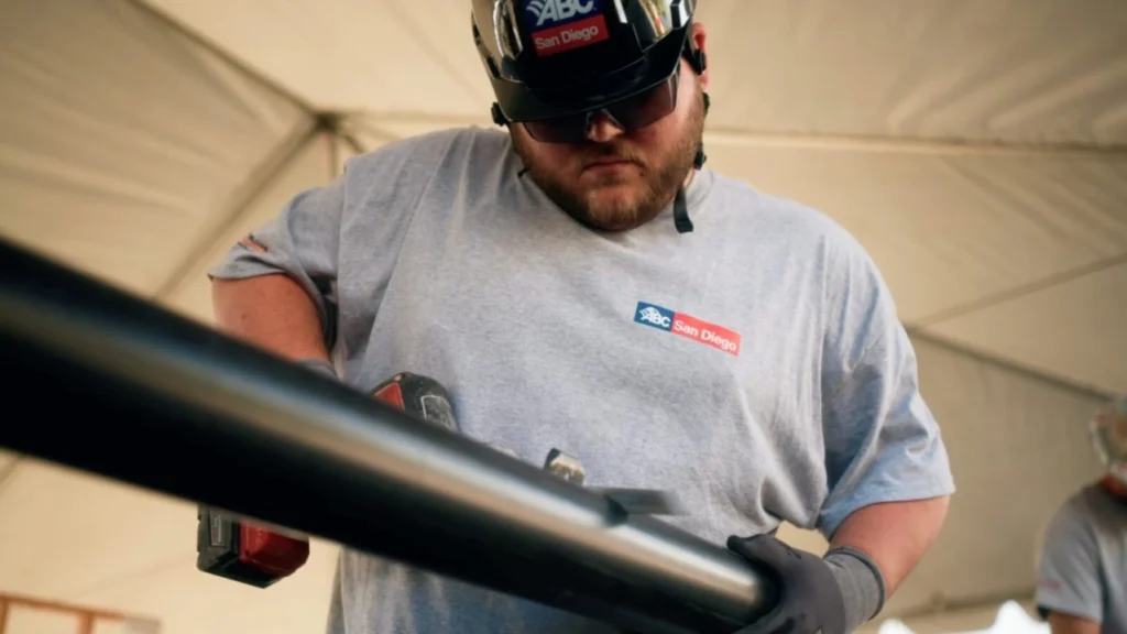 A construction worker wearing safety glasses and an ABC San Diego branded hard hat and t-shirt operating a power tool on a pipe inside a work tent.